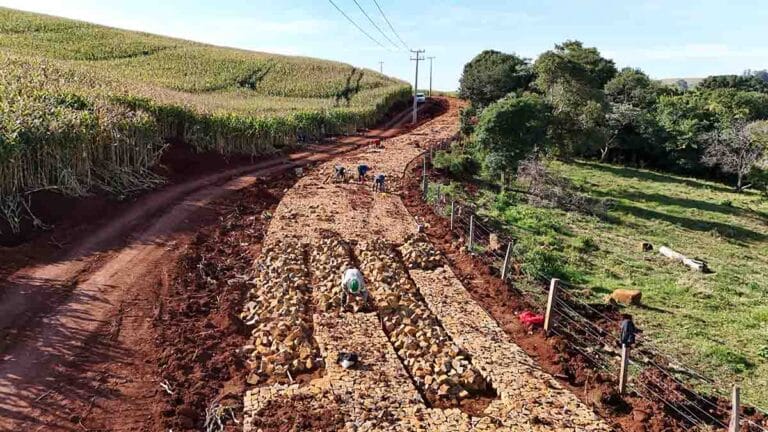 Obra de pavimentação com pedra irregular em estrada rural cercada por lavoura